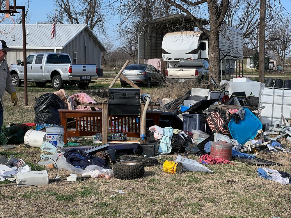 A person stands near a large pile of debris and discarded household items scattered across a yard after a storm.