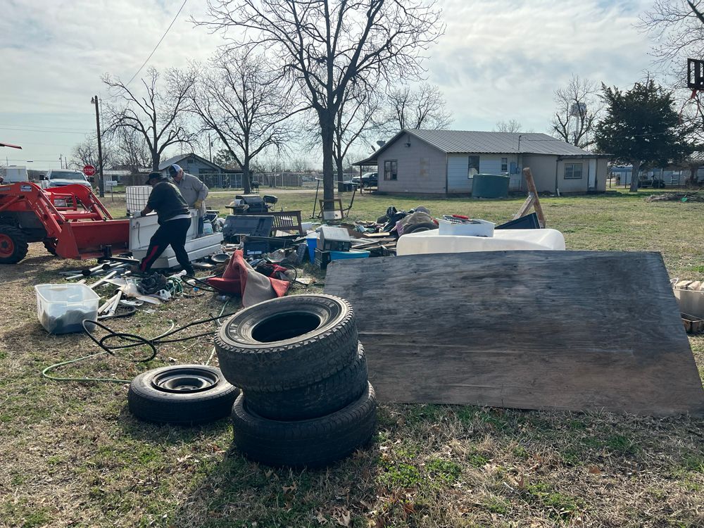 Two people sort through debris and stacked tires in a grassy yard near a house, aided by an orange tractor.