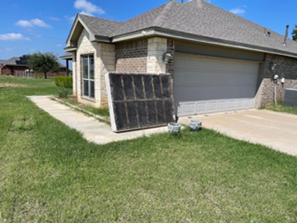 A large, rectangular solar collector panel leans against the stone exterior of a suburban house near the garage driveway.
