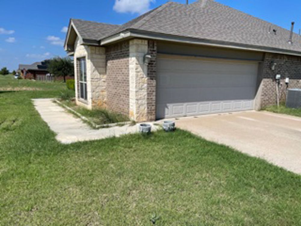 A tan brick and stone house with a two-car garage, a concrete driveway, and a front walkway under a clear blue sky.