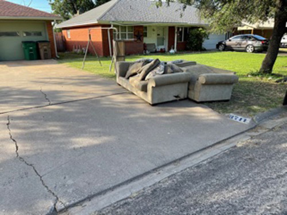 A tan sofa and armchair sit on a concrete driveway in front of a brick house with a green lawn.