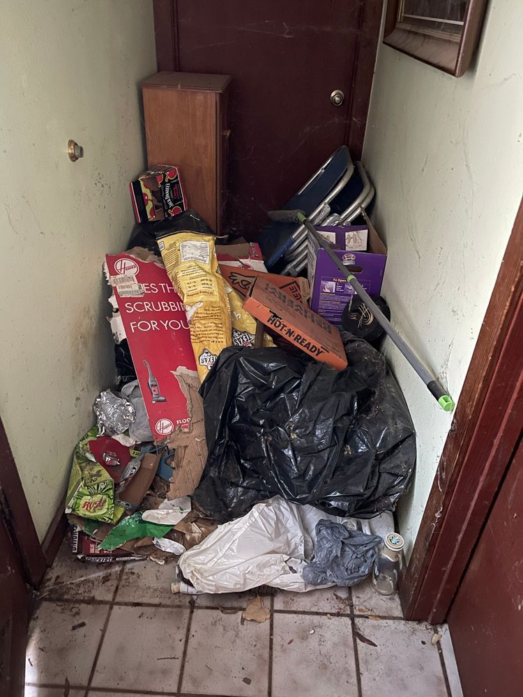 Cluttered, narrow indoor hallway filled with trash bags, boxes, a chair, and miscellaneous items on a tiled floor.