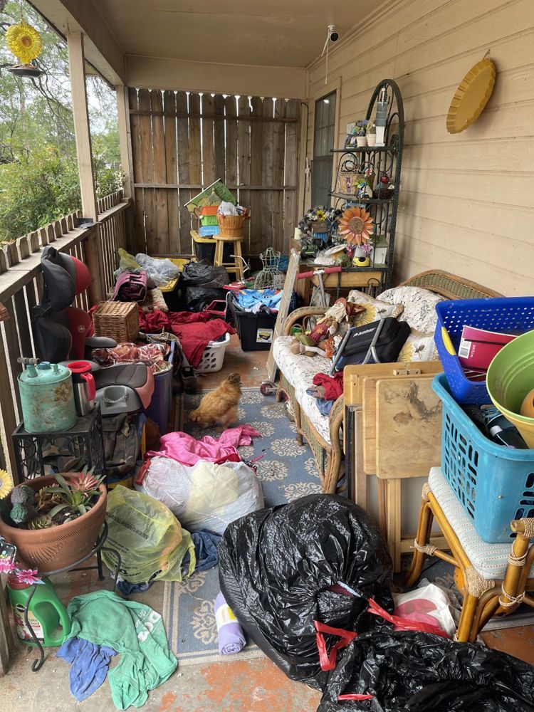 A cluttered covered porch with bags, baskets, furniture, and a small dog sitting on a patterned rug.