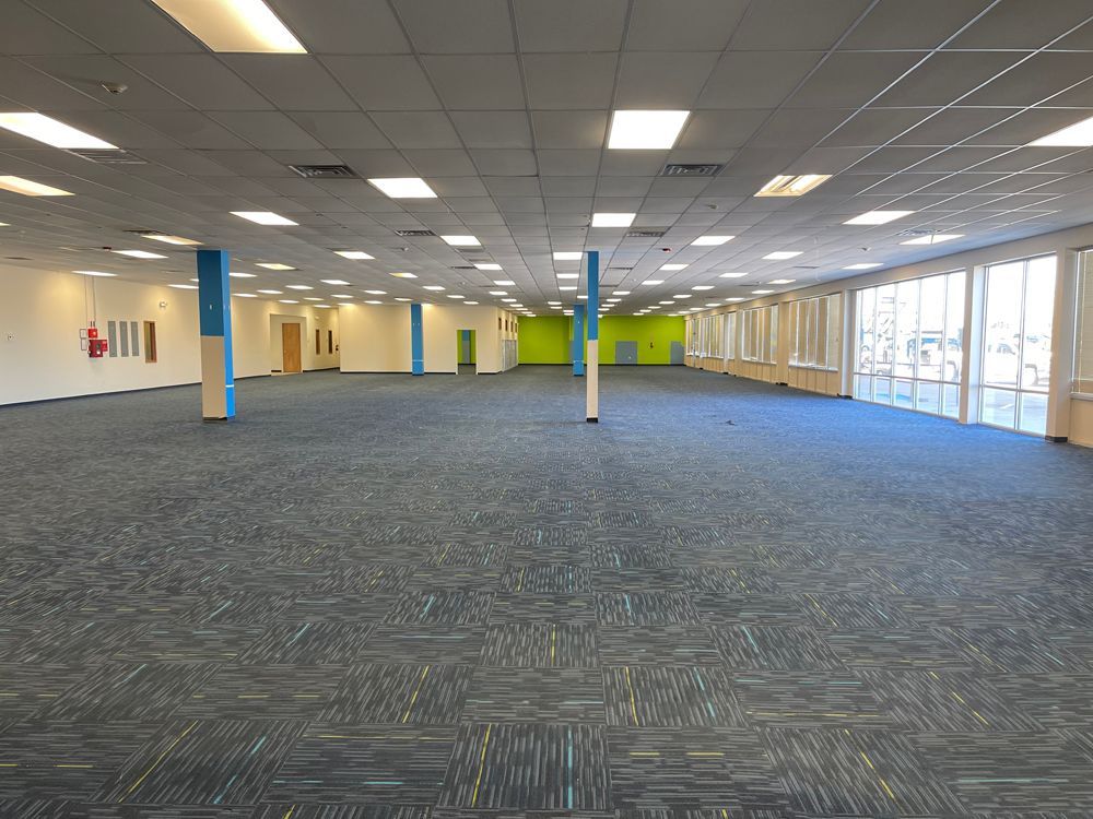 A vast, empty office space with blue patterned carpeting, white walls, and a dropped ceiling with recessed lighting.