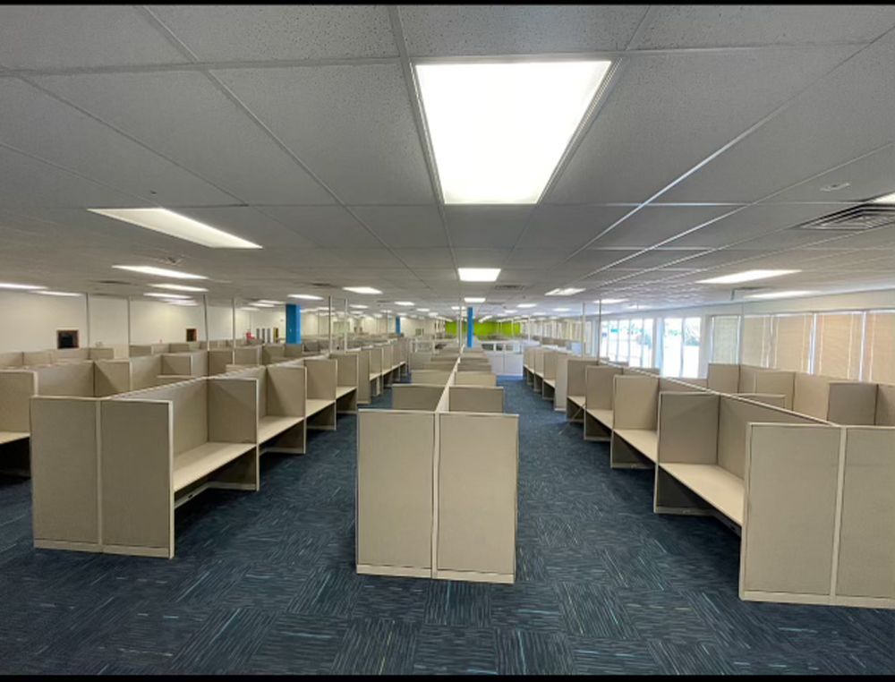 An empty office space with rows of tan cubicles and blue patterned carpet under bright overhead fluorescent lights.