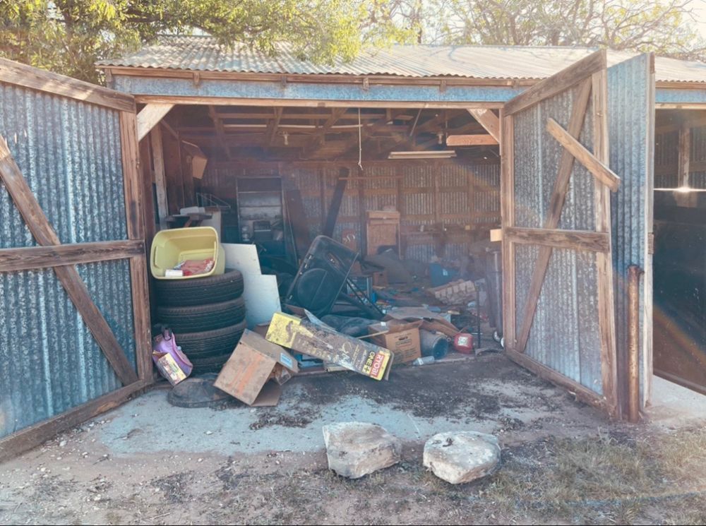 A cluttered, open-doored shed with metal walls containing tires, boxes, and miscellaneous equipment.