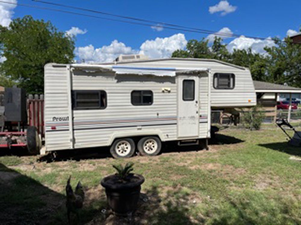 A fifth-wheel camper sits on a grassy lawn under a blue sky, with a dark pot and a chicken in the foreground.