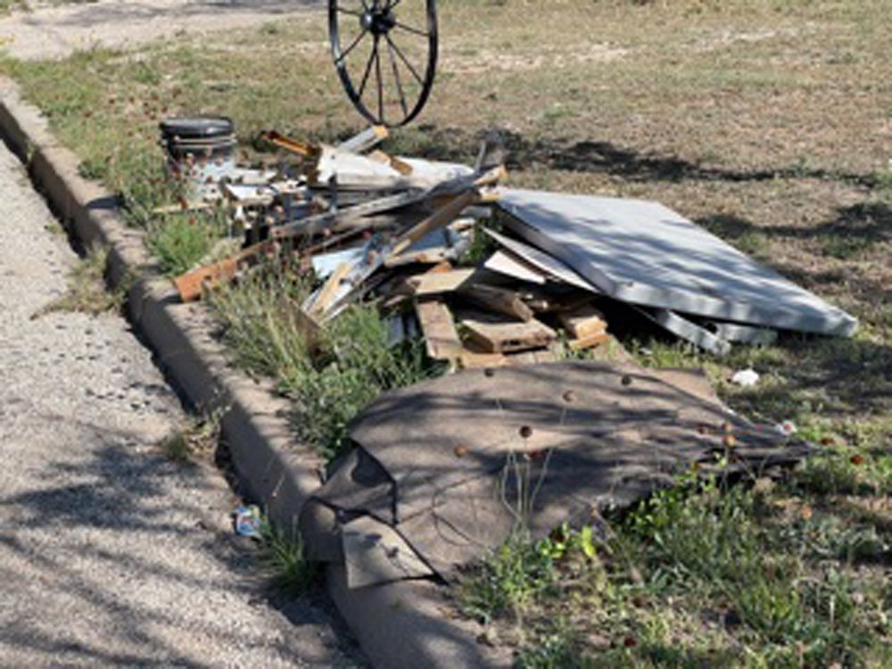 A pile of discarded debris, including wood scraps, a white board, a bucket, and a metal wheel, sits on a grassy curb.