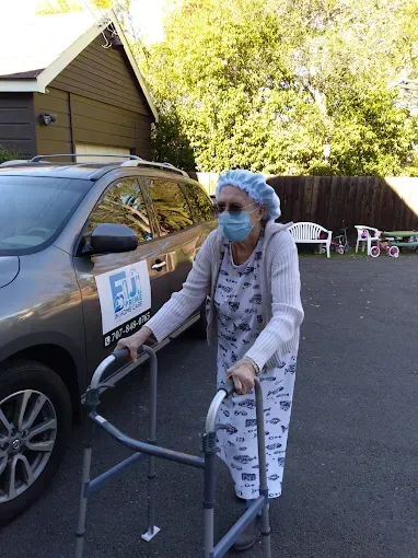 Person using a walker, wearing a mask and hair covering, next to a car.