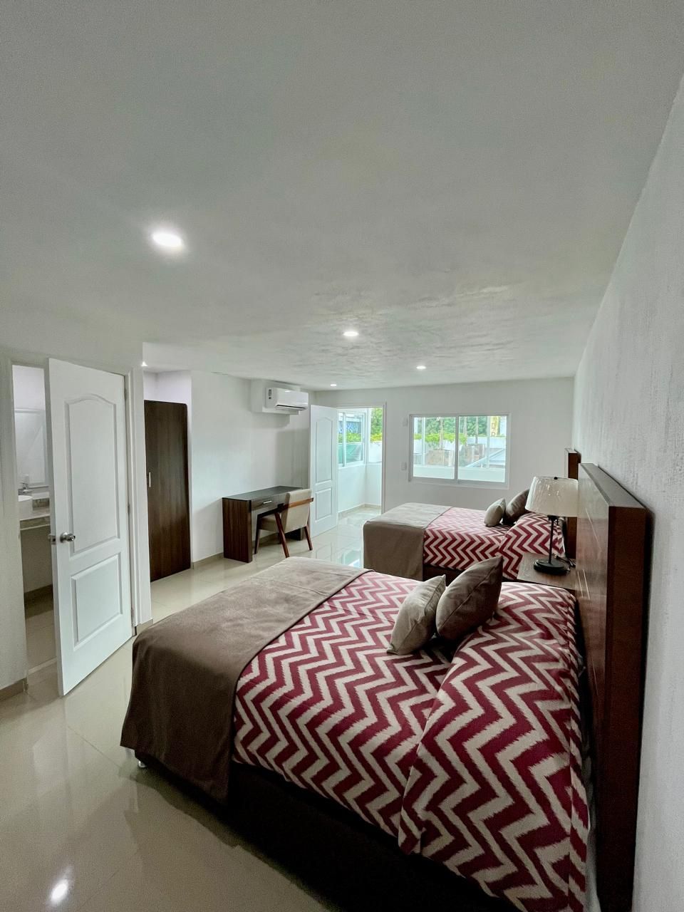Habitación de hotel con dos camas, escritorio y puerta abierta al baño. Habitación luminosa con paredes blancas