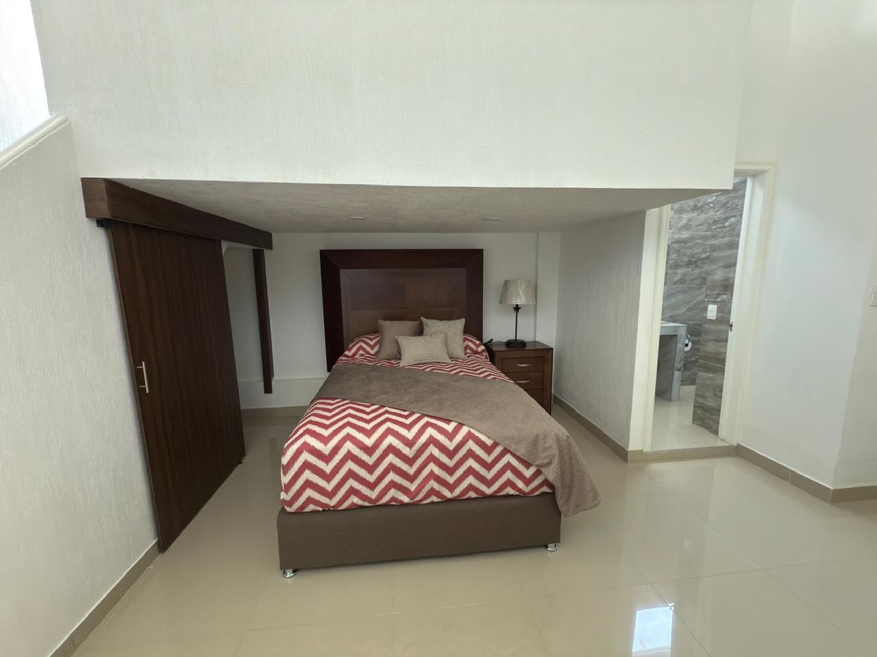 Dormitorio con cama bajo un altillo, cabecero marrón, manta de chevron rojo y blanco, puerta a la izquierda 