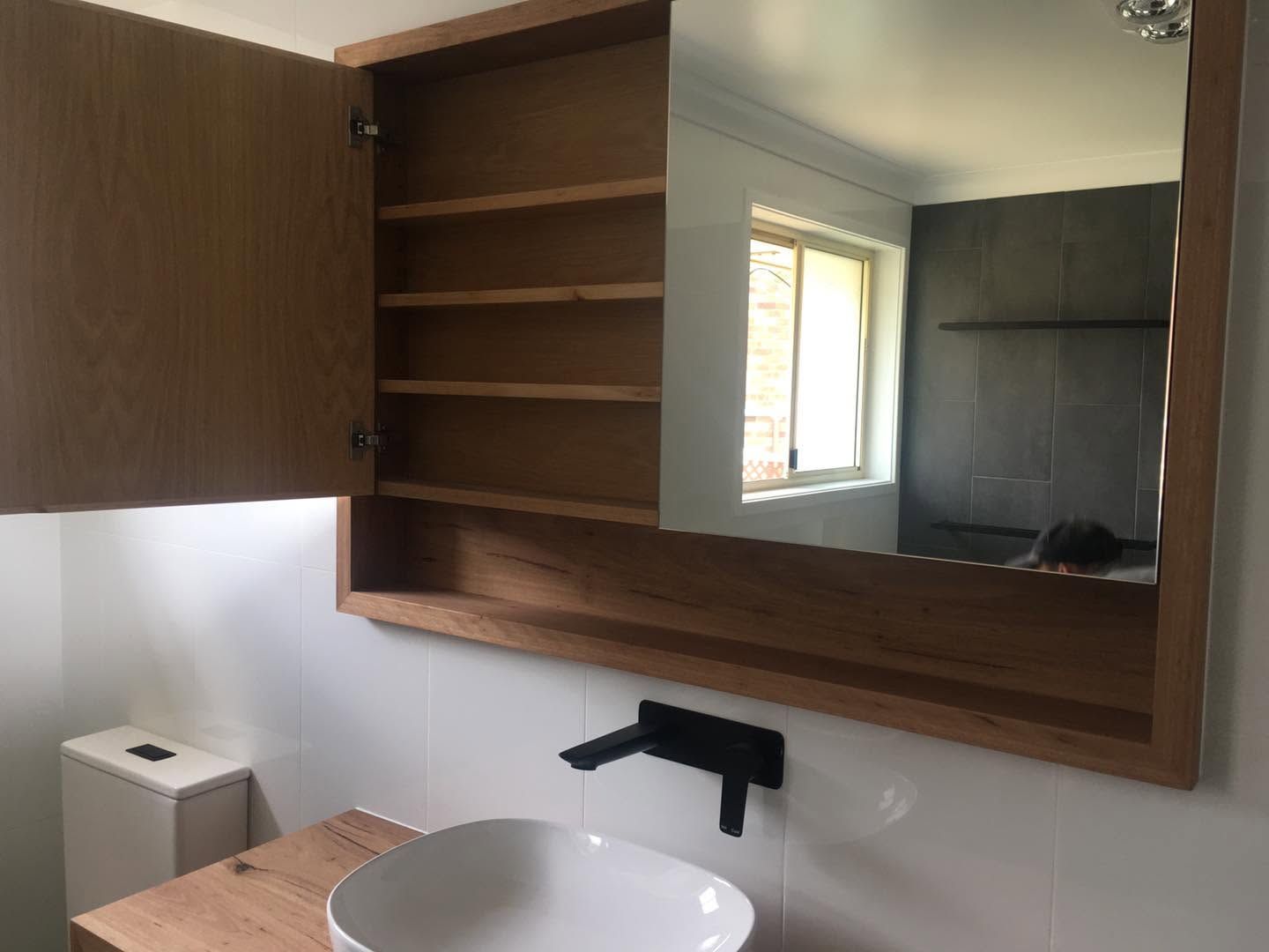 Bathroom vanity with open wood cabinet, mirror, sink, and black faucet beside a window.