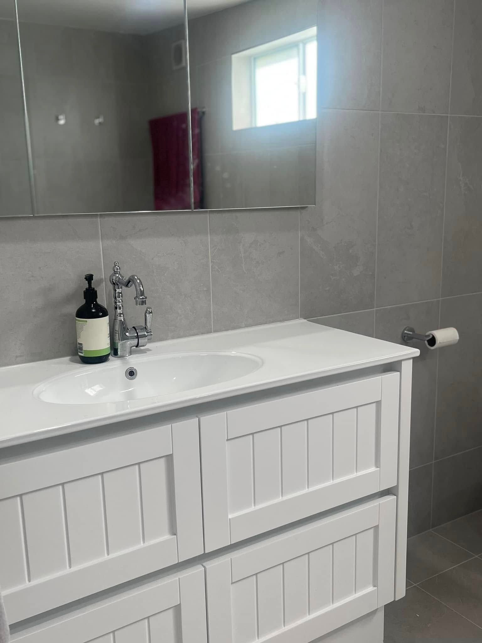 Modern bathroom vanity with white sink cabinet, soap dispenser, and gray tiled wall with mirror and window.