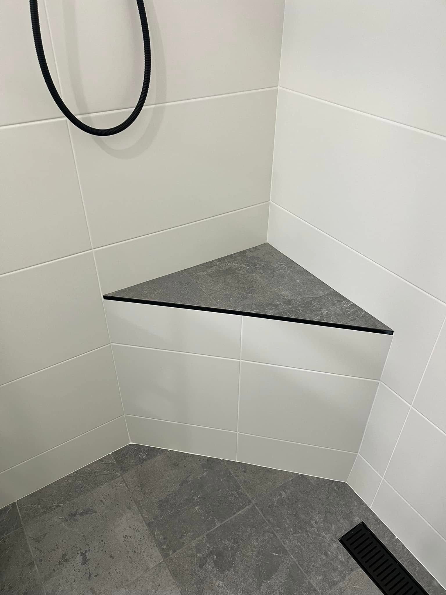 Corner shower with white tile, gray bench, and dark shower floor drain.