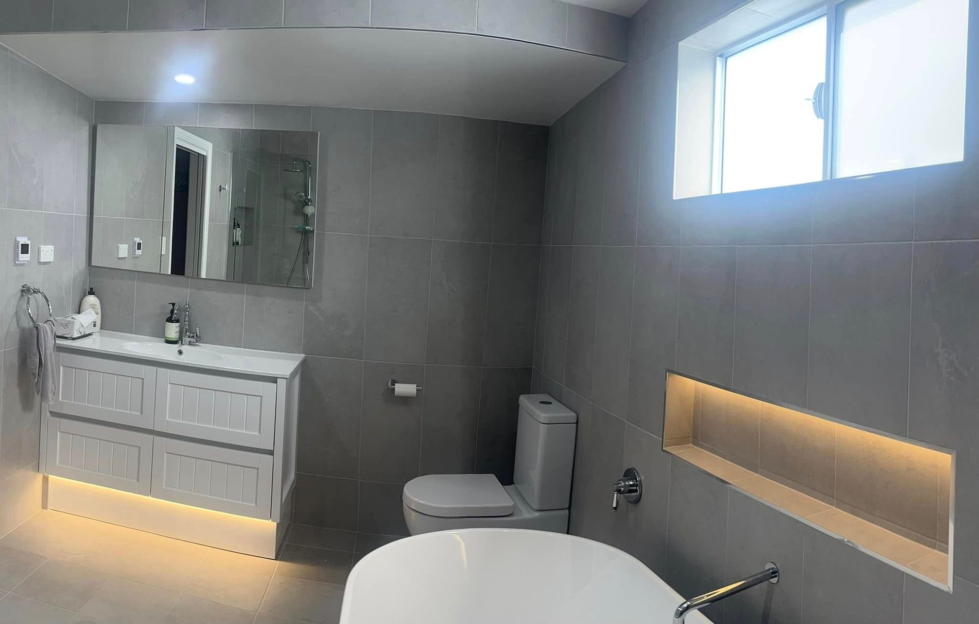 Modern gray bathroom with vanity, toilet, tub, recessed wall niche, and a window.