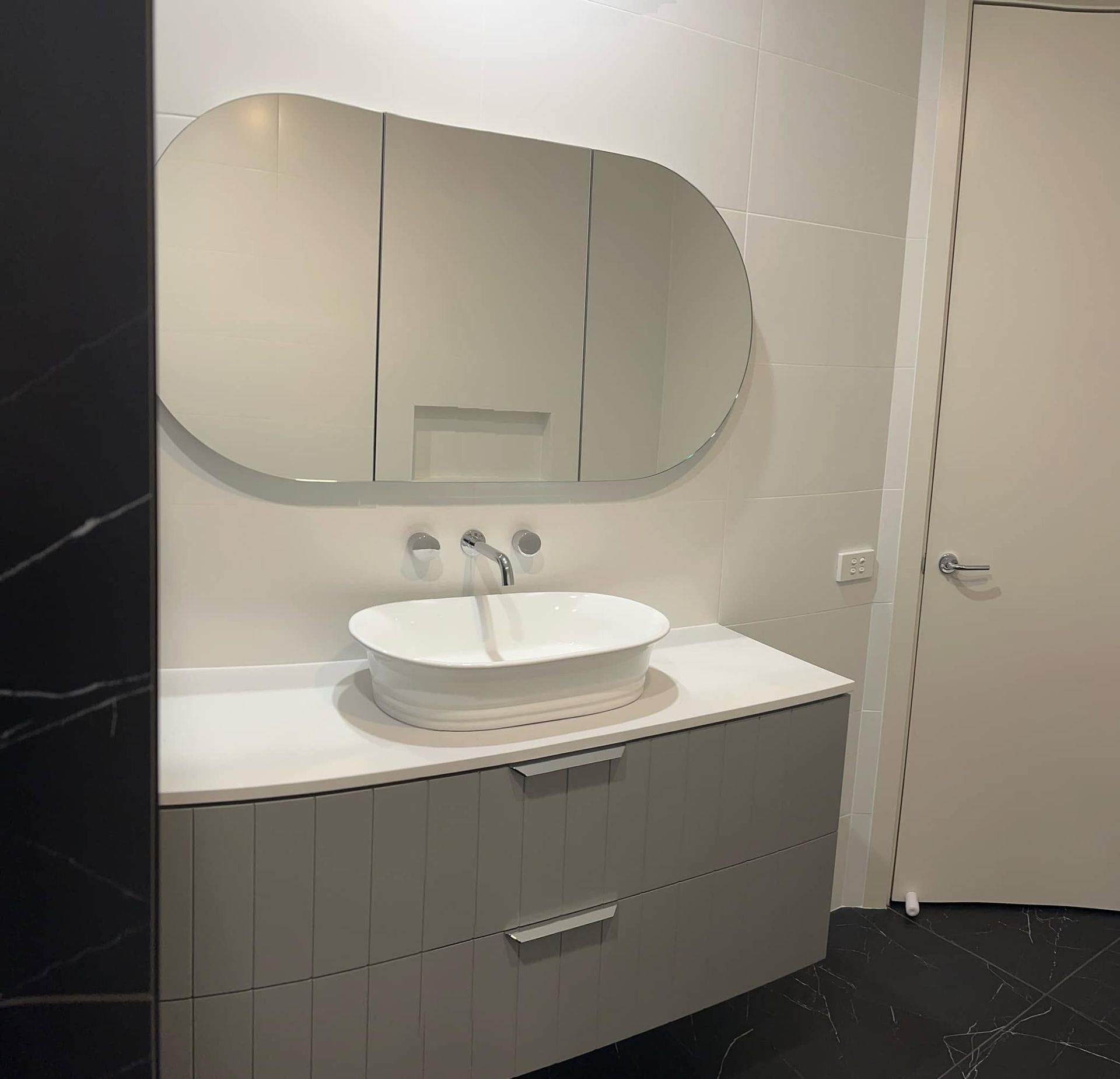 Modern bathroom vanity with oval mirror, white sink, and gray cabinet beside a closed door