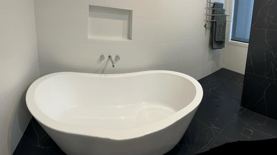 Modern white freestanding bathtub in a minimalist bathroom with dark floor tiles and wall-mounted faucets