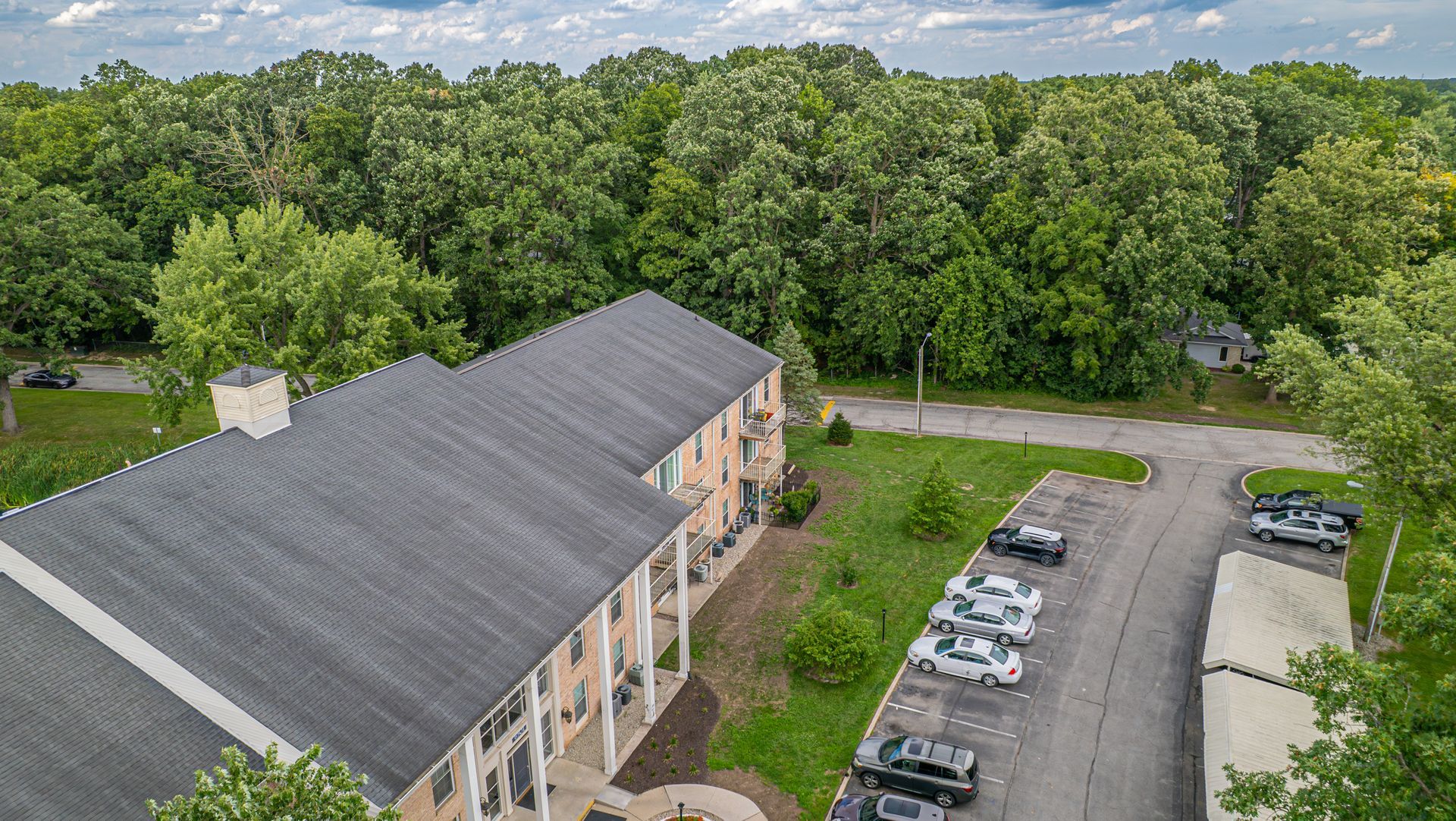 Two-story building with a gray roof, surrounded by trees and a parking lot with cars.