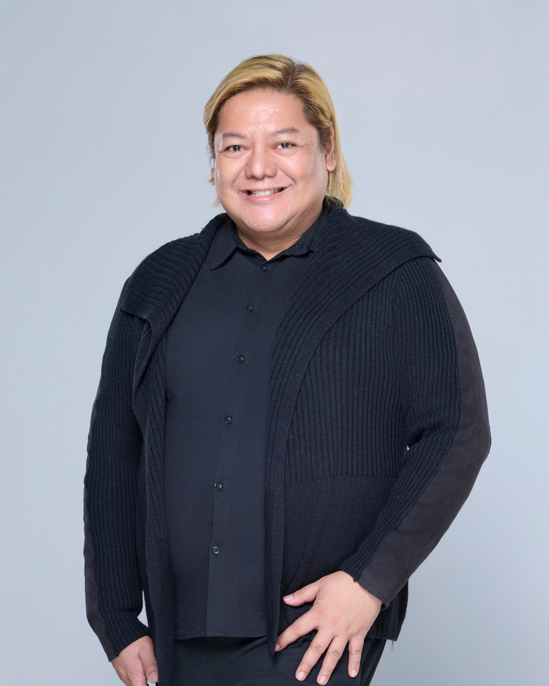 Man with blonde hair, smiling, wearing a black jacket and shirt, standing in front of a white background.