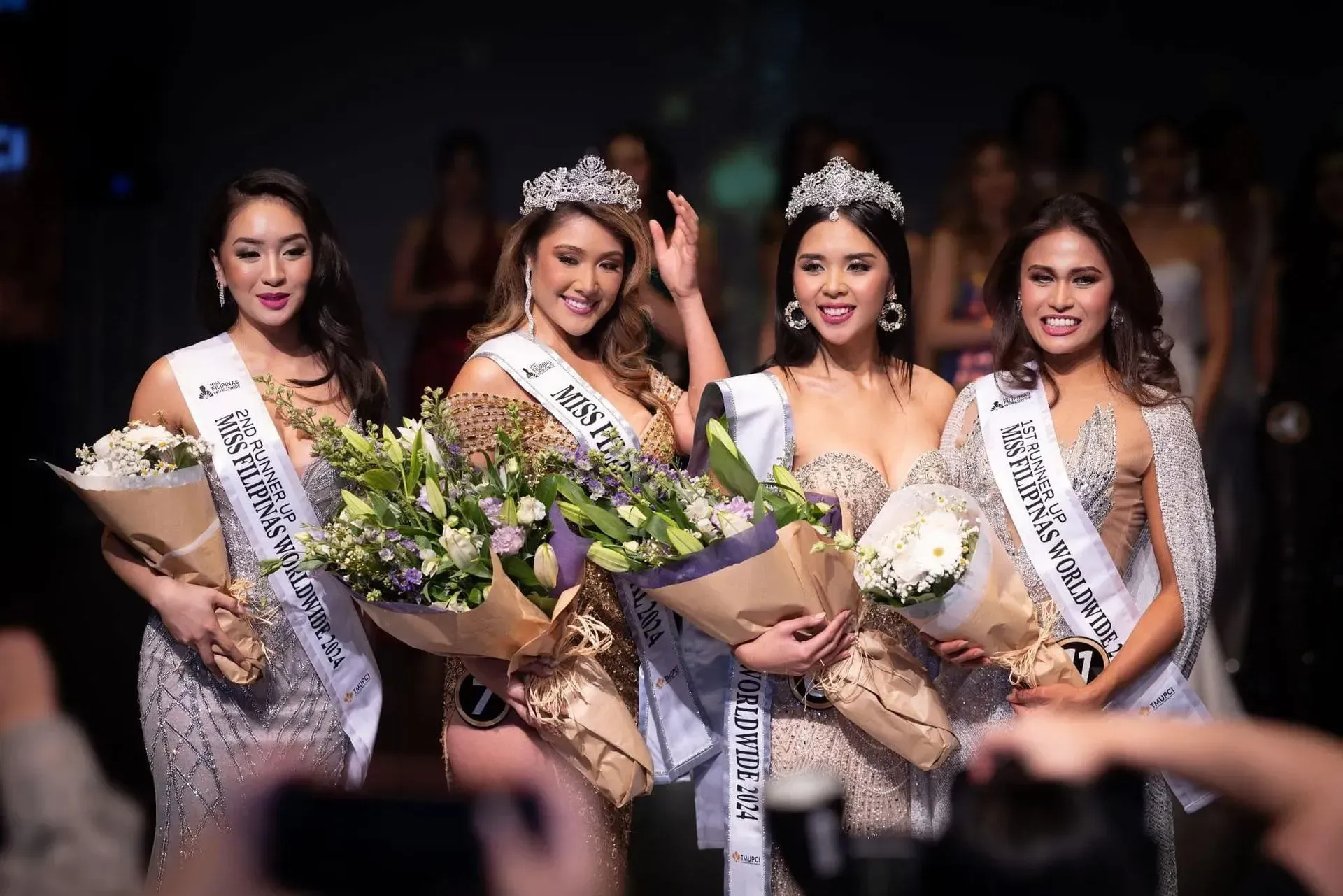Four beauty queens in crowns and sashes holding bouquets, smiling, on a stage.