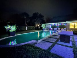 Backyard pool lit with blue-green lights at night, beside a covered patio and stepping stones