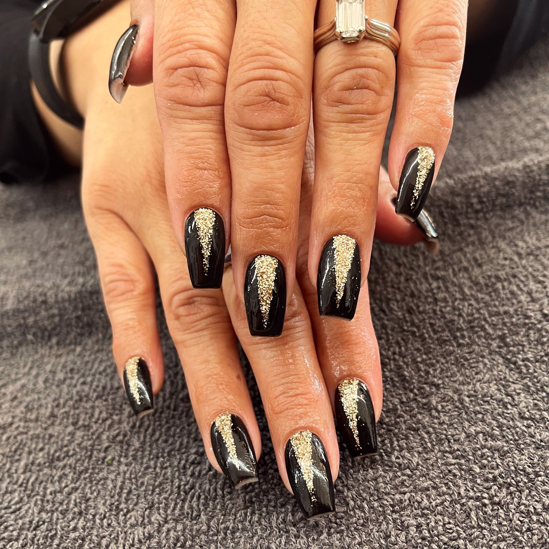 Hands with long black glittery acrylic nails resting on a gray surface.