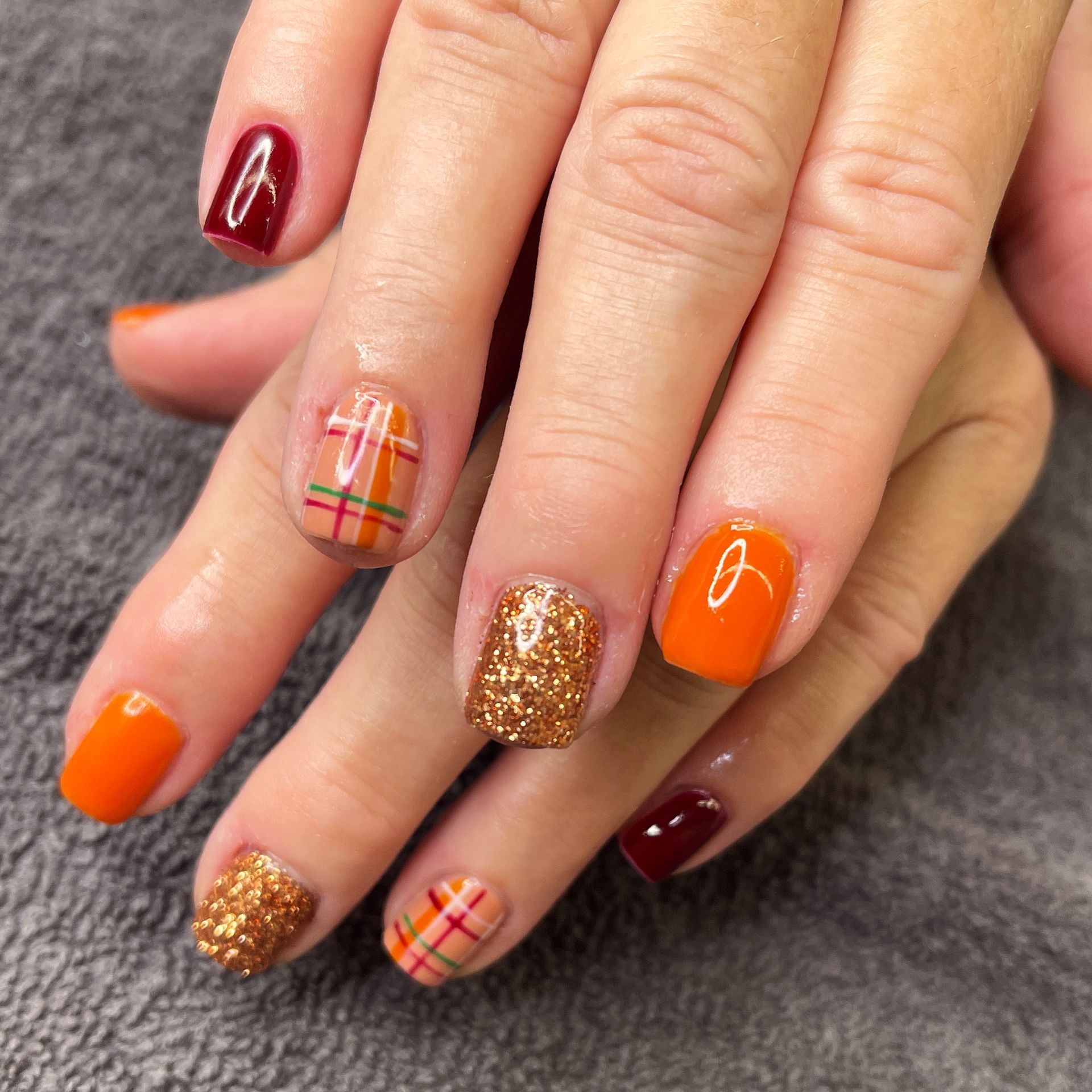 Close-up of manicured hands with orange, burgundy, gold glitter, and plaid nail art