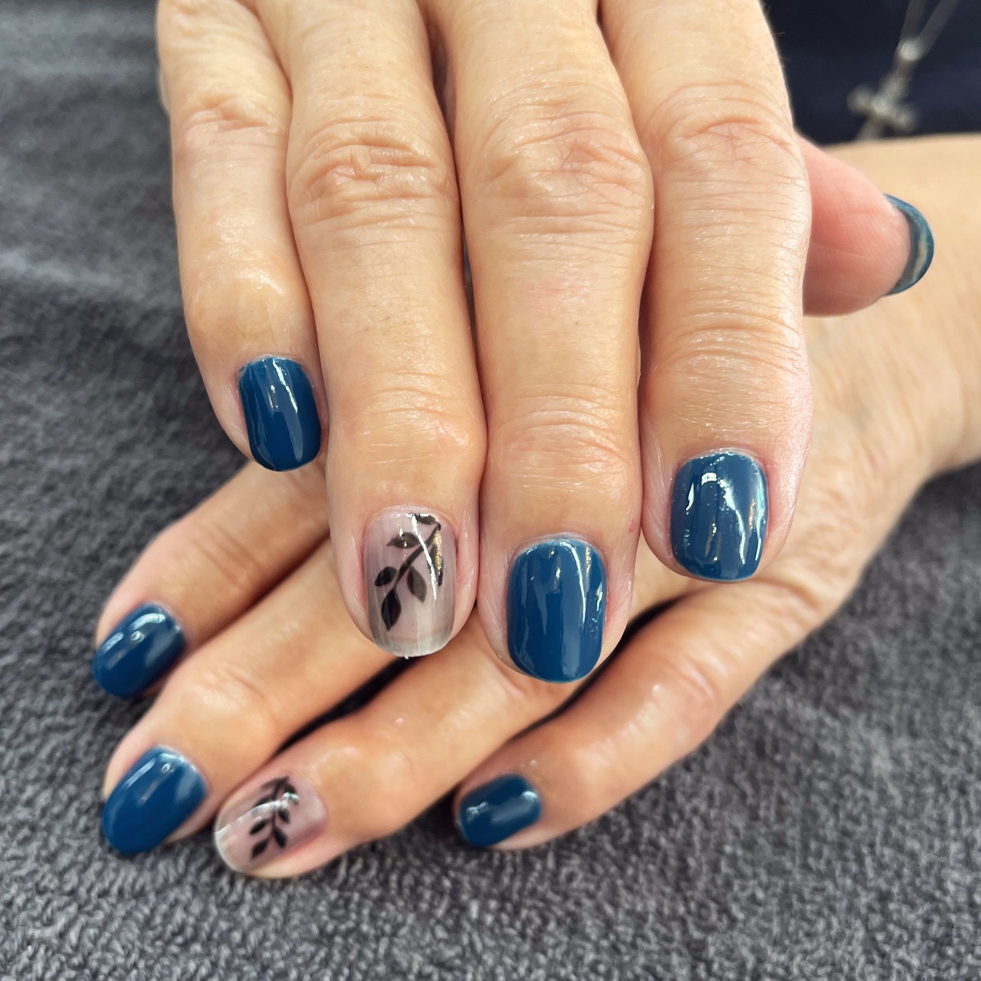 Hands with glossy teal nails and two nude accent nails with black leaf designs.