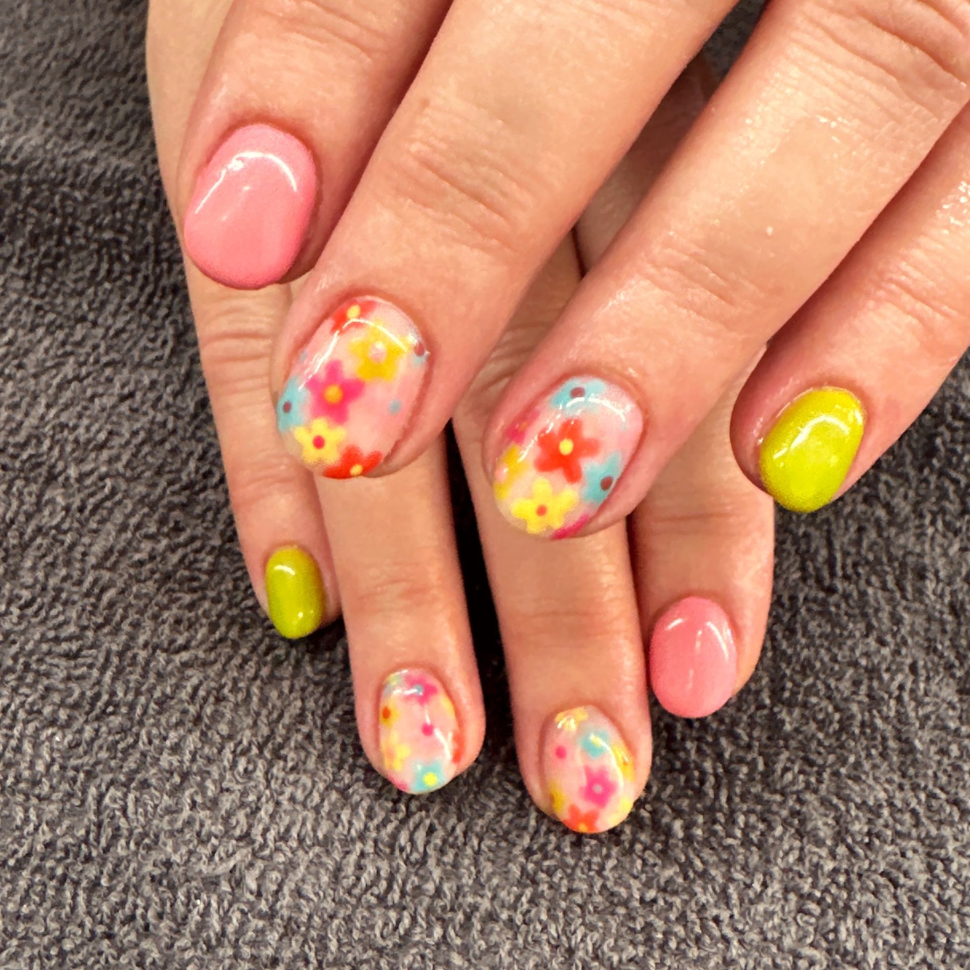 Hands with colorful floral nail art on pink and neon yellow nails over a gray towel