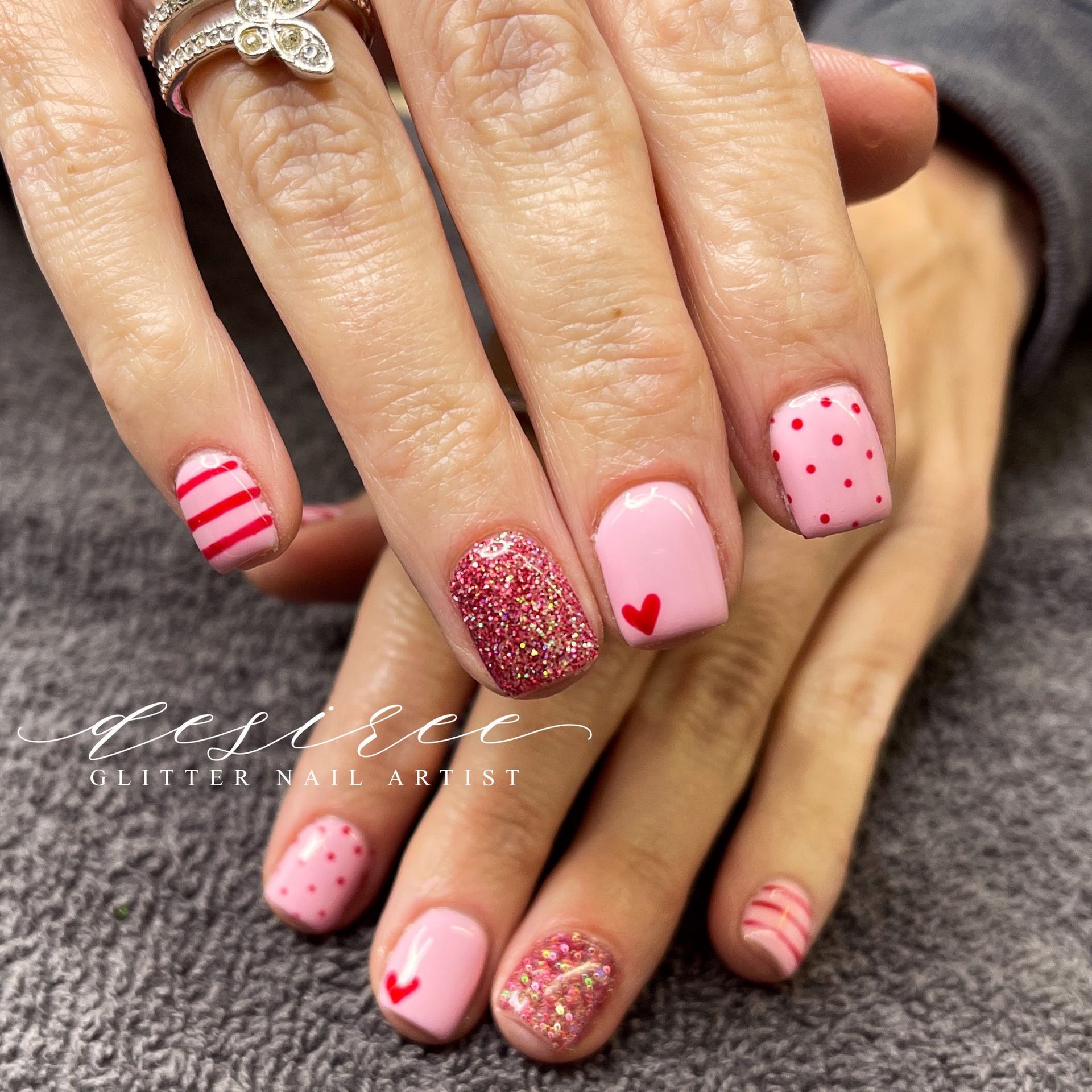Hands with pink Valentine-themed nail art, including hearts, dots, stripes, and glitter.