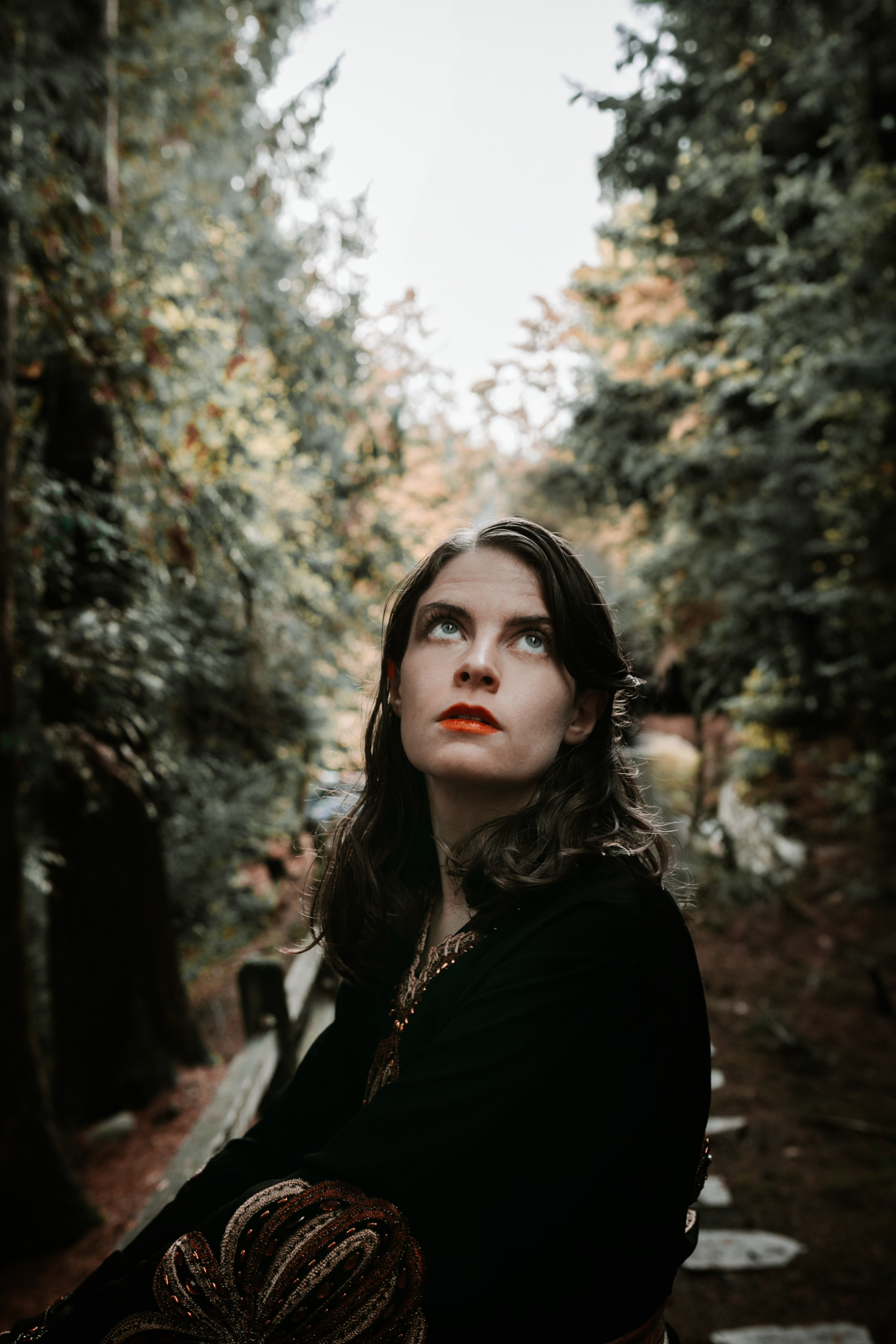 Woman looking upwards in a forest, wearing black and a red lip. Trees surround her.