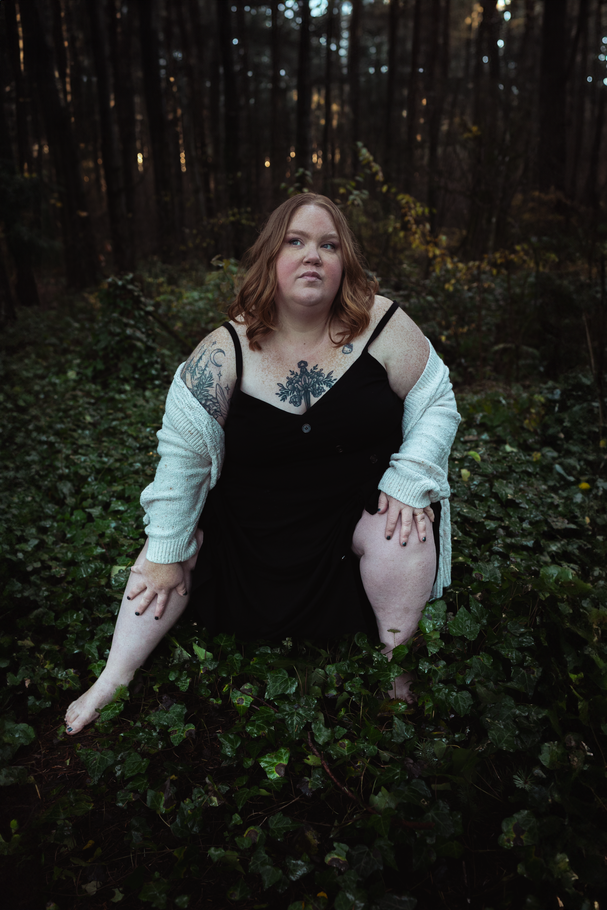 Plus size woman sits among green foliage in a dark forest, wearing a black dress and white cardigan.