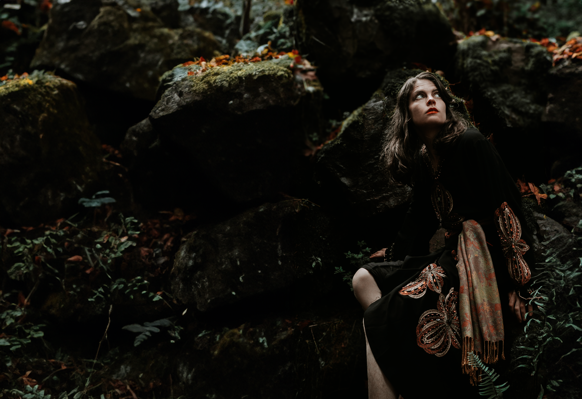 Woman in dark clothing looks up, posed against mossy rocks and foliage.