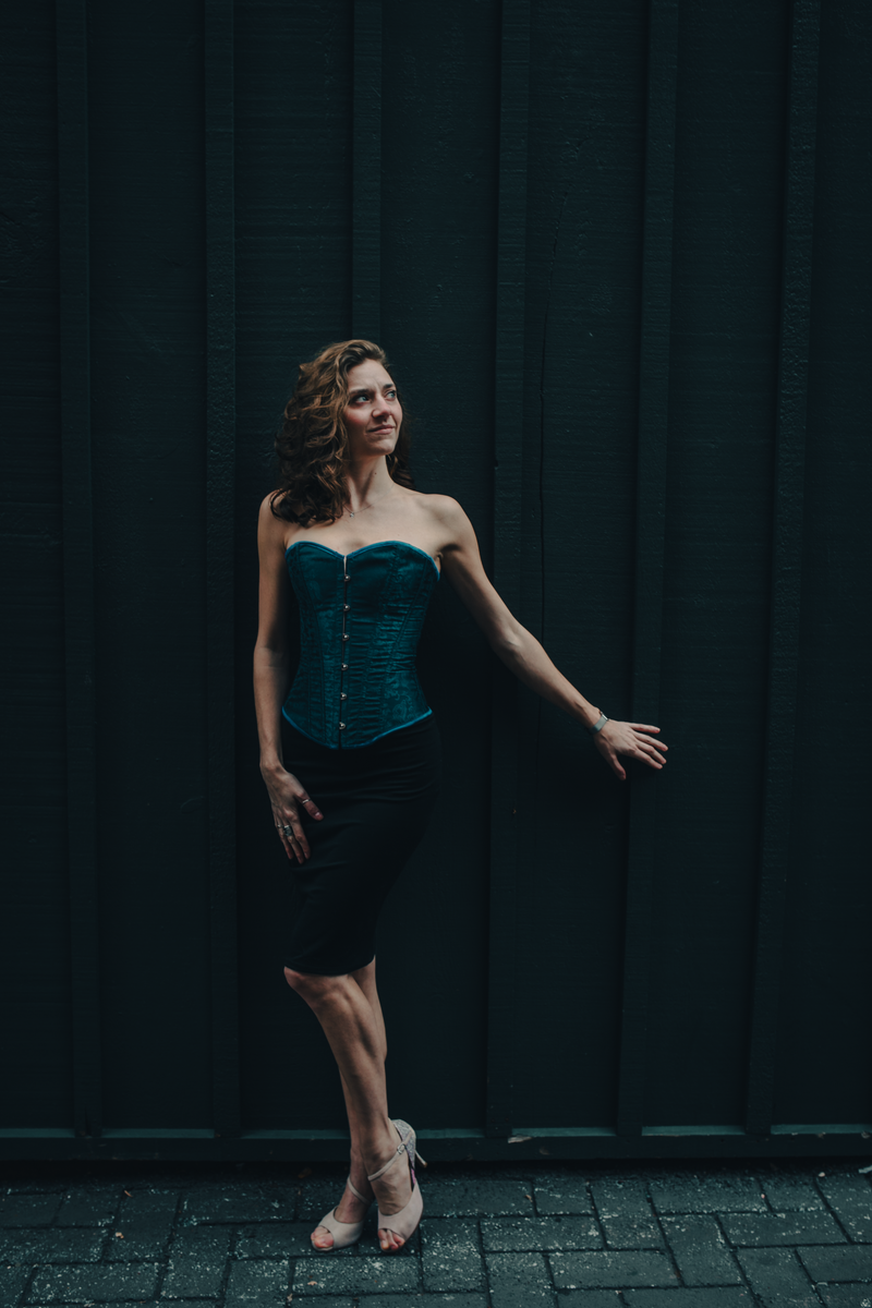 Woman in teal corset and black skirt, poses against a dark wall, looking upward, arm outstretched.