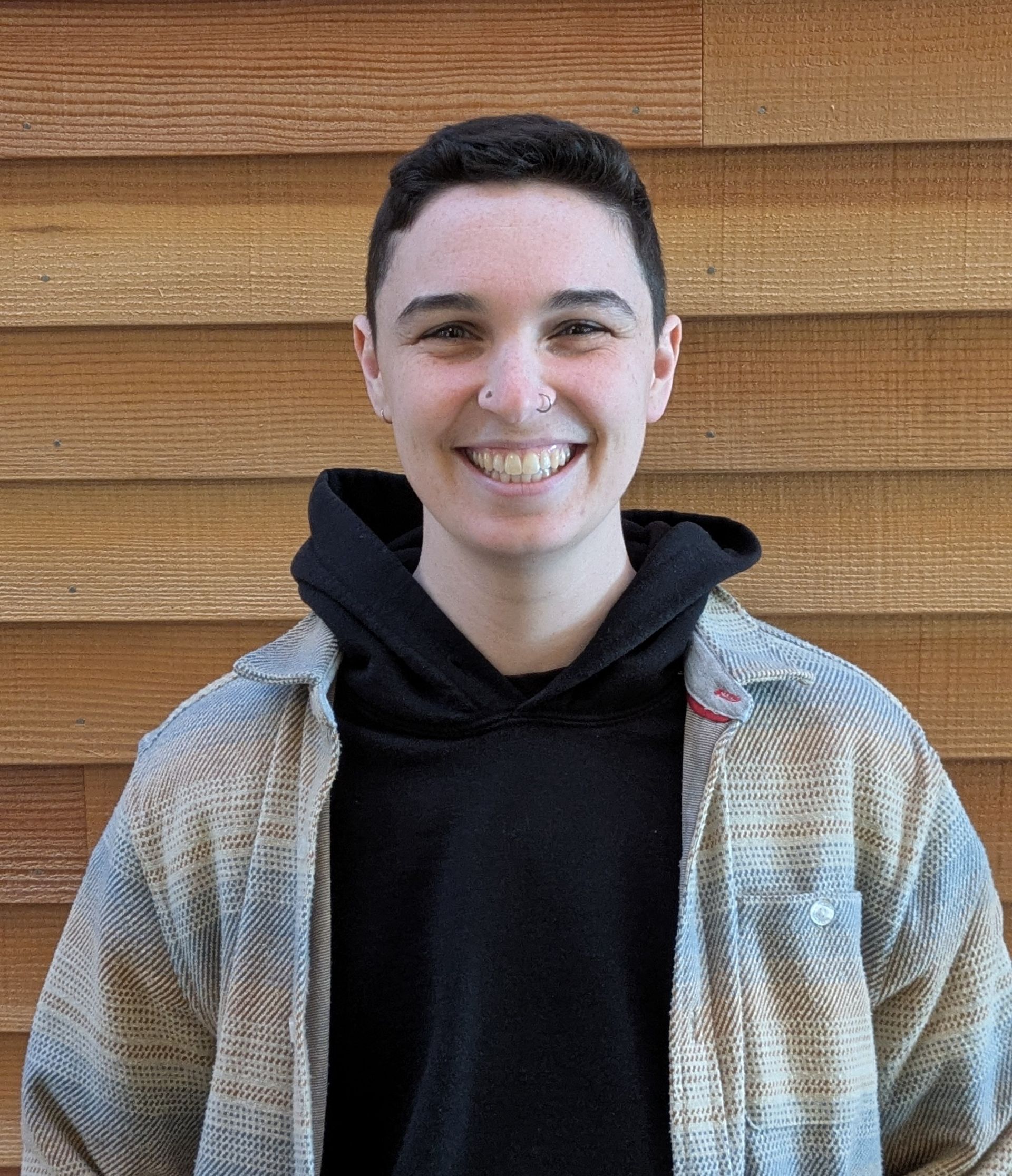 Person with glasses smiles, wearing a light blue flannel over a black hoodie, in front of a wood-paneled wall.