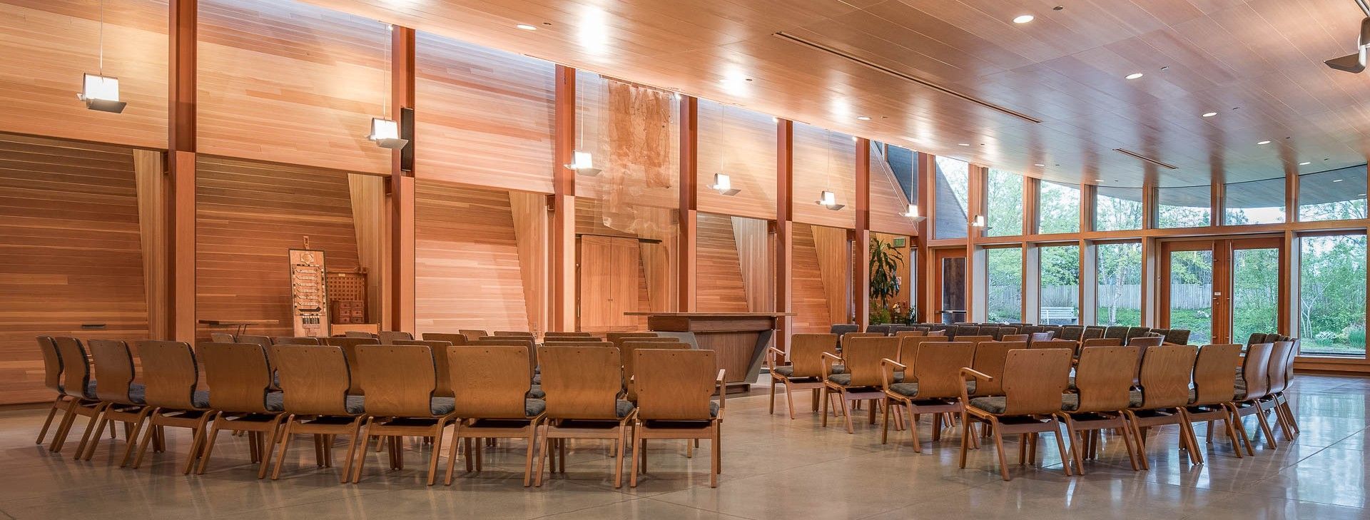 A wooden chapel with rows of chairs in a circle. Large windows and wood paneling provide natural light.