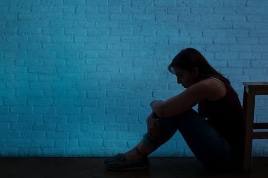 Silhouetted person sitting on the floor against a blue brick wall, knees hugged in a dim room.