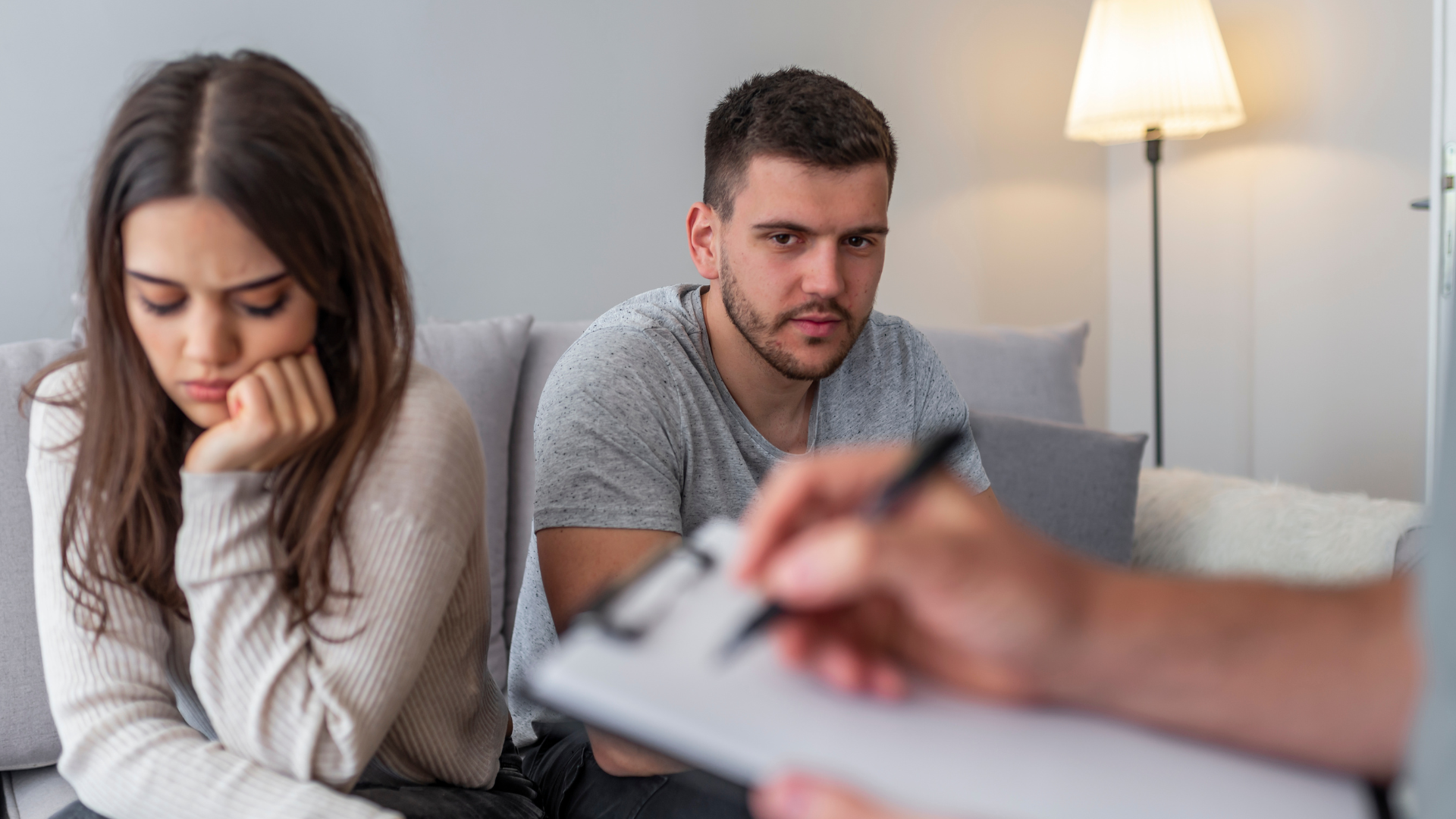 Couple in counseling, woman upset on sofa while man listens as therapist takes notes