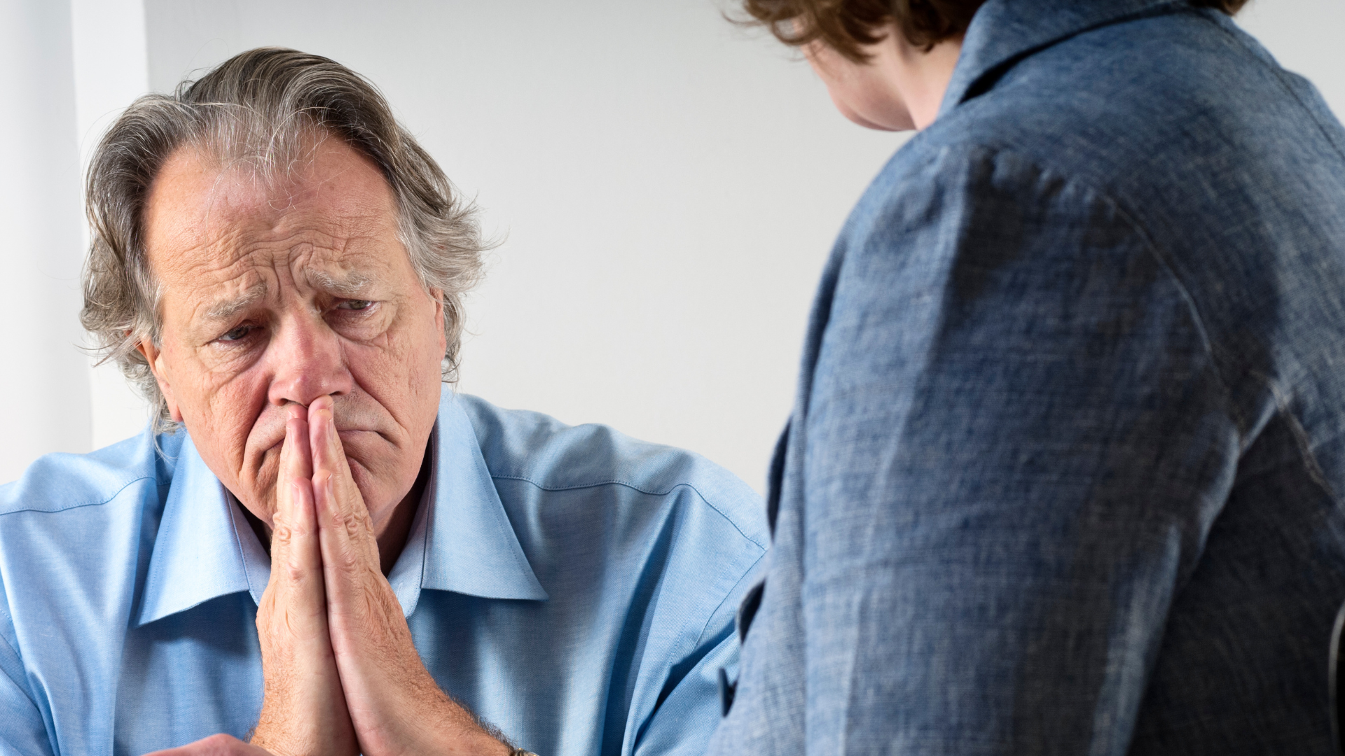 Man in blue shirt with clasped hands, looking distressed while talking to another person