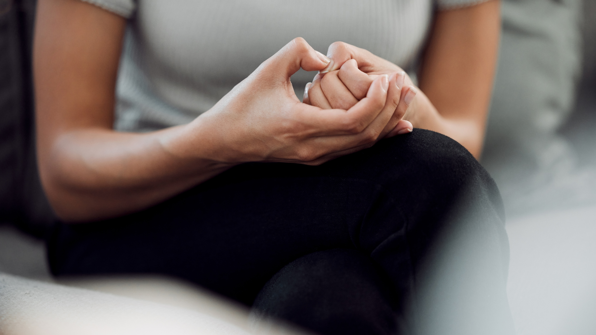 Close-up of hands clasped over a bent knee, wearing a gray top and black pants, seated on a couch.