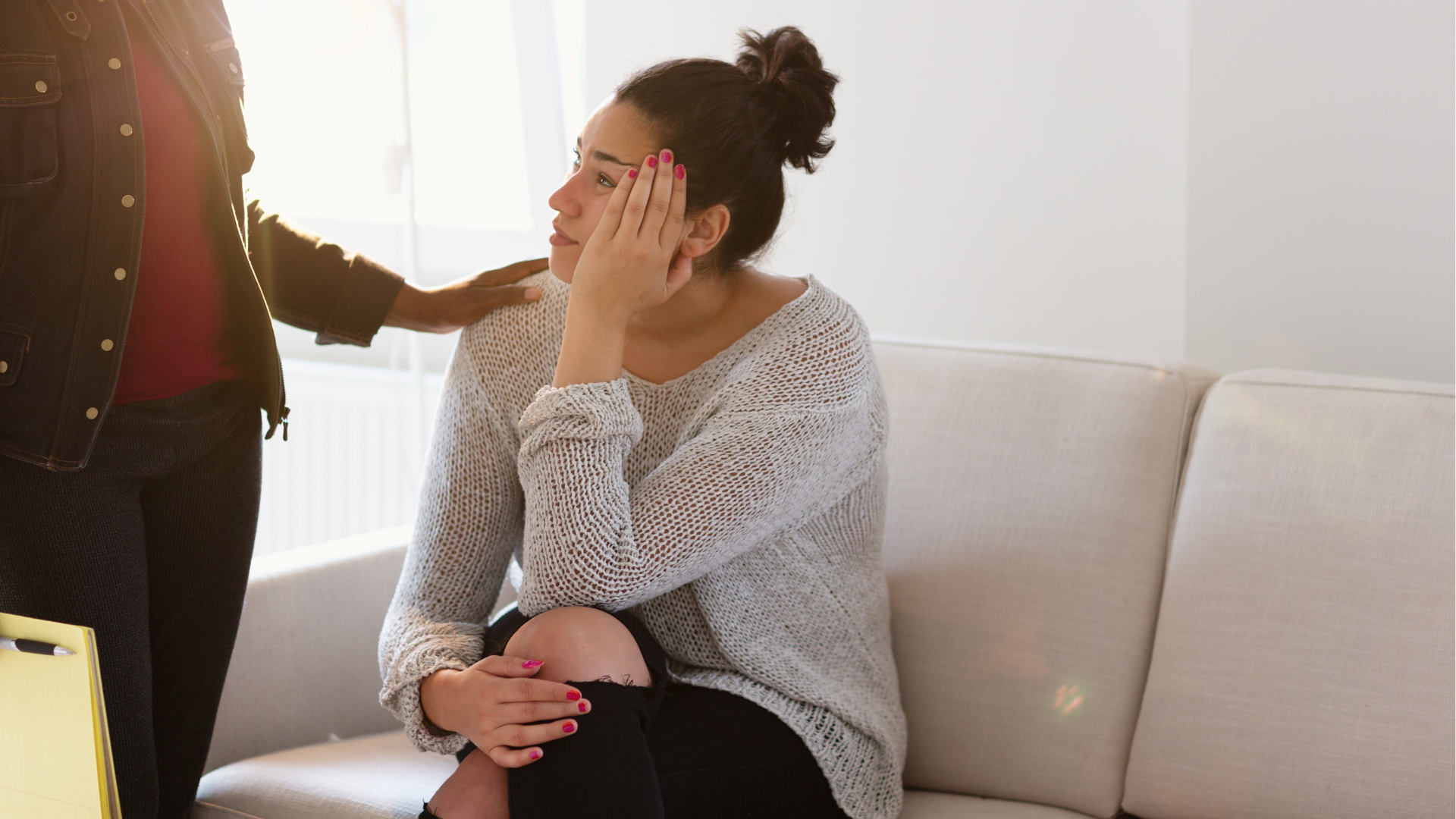 Person sits on a couch, covering their face while another person stands nearby with a comforting hand on their shoulder.