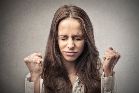 Woman with eyes closed, clenching fists, showing frustration against a plain background