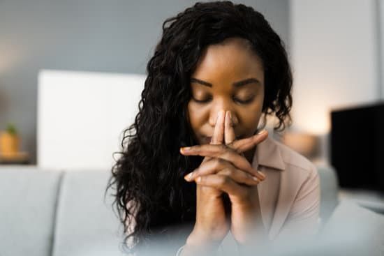 Person seated at a desk, hands clasped near face, looking down in a thoughtful pose in a bright office.