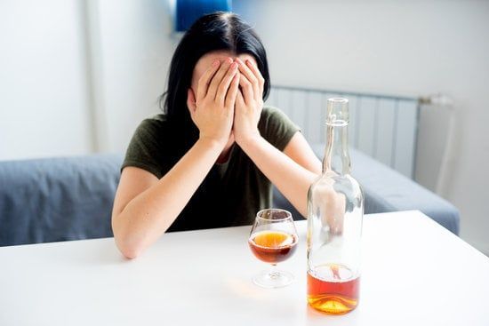 Person at a table with face in hands beside a glass and bottle of amber liquid