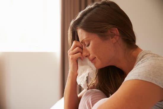 Woman sitting by a bright window, covering her eyes with a tissue and looking upset