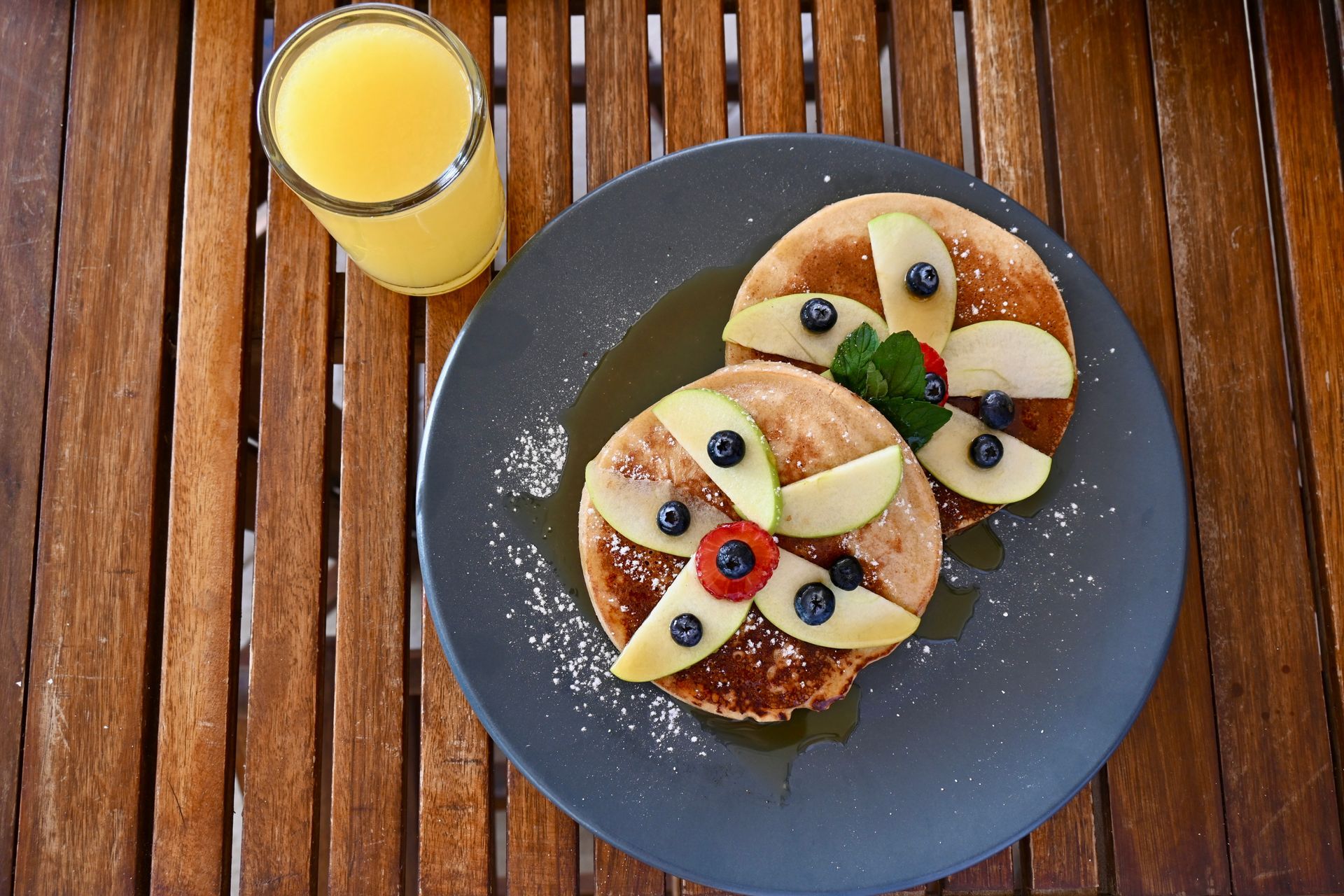Un plato de panqueques con fruta y un vaso de jugo de naranja en una mesa de madera.
