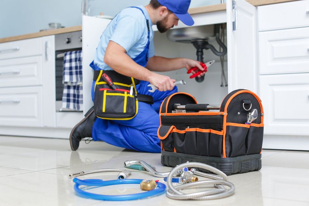 Plumber in blue overalls kneeling under a sink, working with tools from a toolbox in a kitchen.