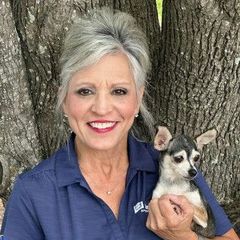 Woman with gray hair smiling, holding small dog in front of a tree.