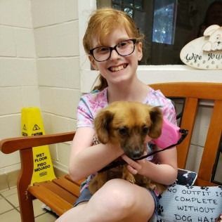 Girl with glasses smiles, holding a small brown dog on a bench, inside a room with a 