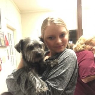 Blonde woman holding a small gray dog. Another person is visible in the background.